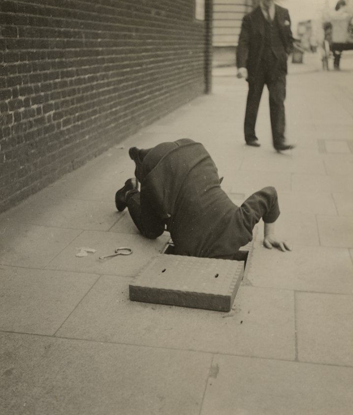 Black and white photograph of a man looking inside a sidewalk inspection door in London pavement